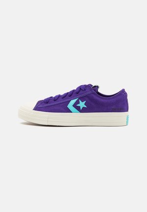 Fioletowy niski sneaker z turkusową gwiazdą i logo w kształcie jodełki, białą gumową podeszwą i sznurowadłami, widziany z boku.