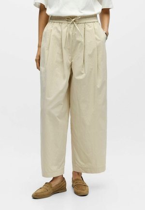 Personne portant un pantalon beige ample court avec taille à cordon et mocassins en daim marron clair, debout devant un fond uni.