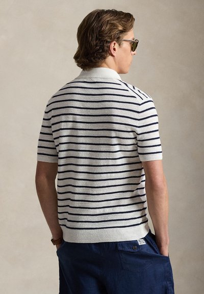 Short-sleeve polo shirt in cream with navy stripes, featuring a ribbed collar and cuffs. Worn with navy shorts and sunglasses.