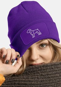 Purple knitted beanie with a ribbed texture featuring a white dog silhouette on a patch. The beanie is worn with a brown scarf.