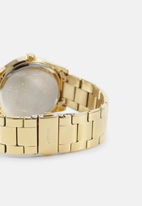 Guld rustfrit stål ur med rund urkasse, armbåndsstil og logoindgravering på låsen. Glat, poleret overflade med en reflekterende finish.