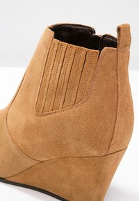 Bottine compensée en daim camel avec des détails de couture verticale, bout arrondi et semelle noire. Texture lisse avec une finition douce.