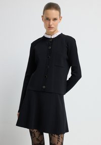 Mustat lyhyet takki kultaisilla napeilla ja etutaskulla, yhdistettynä mustiin hameisiin. Mallilla on päällään kuvioidut mustat sukkahousut.