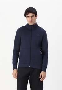 Chaqueta de navy con cremallera y cuello alto, bolsillos frontales, puños acanalados y bajo. Presenta un discreto logo en el pecho. Textura suave.