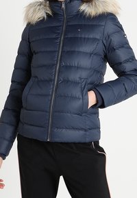 Tmavě modrá péřová bunda s kapucí lemovanou kožešinou, předním zipem a horizontálním prošitím; má přední kapsy a elastické manžety.