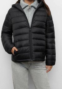 Svart puffjacka med horisontell quiltad mönster, hel dragkedja framtill, huva och sidofickor, bärs över en grå tröja och ljusa jeans.