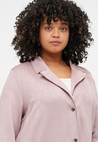 Light pink suede jacket with a notched collar, single button closure, and smooth texture, worn over a white top.