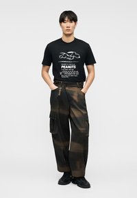 Άντρας που φοράει μαύρο T-shirt με γραφικό χαρακτήρα Peanuts και φαρδιά καφέ και μαύρη cargo παντελόνια, στέκεται με τα χέρια στις τσέπες.