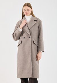 Manteau croisé en tissu texturé beige clair, doté d'un col cranté, de deux poches avant et de boutons noirs.