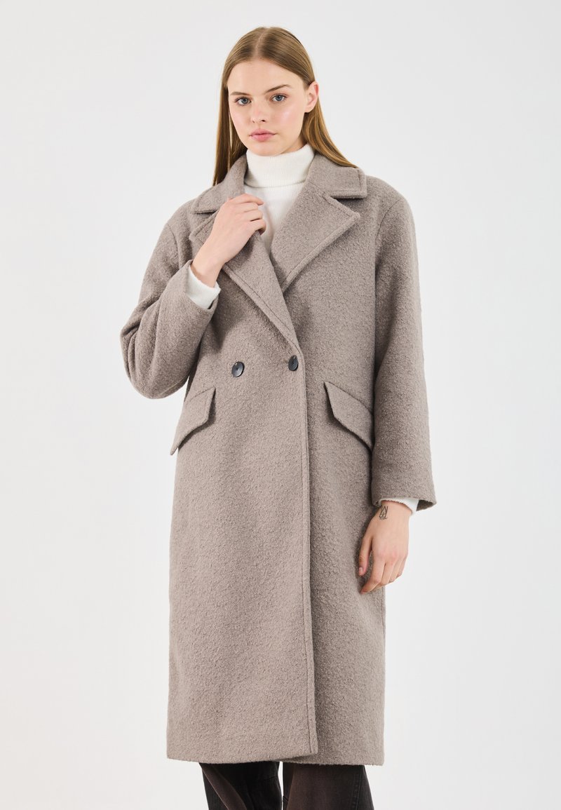 Manteau croisé en tissu texturé beige clair, doté d'un col cranté, de deux poches avant et de boutons noirs.