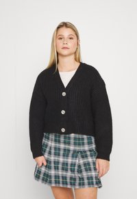 Černý kratší cardigan s žebrovanou texturou a pěti knoflíky, v kombinaci se sukní s plisováním v zeleno-bílé kostkované vzoru. Měkká pletenina.