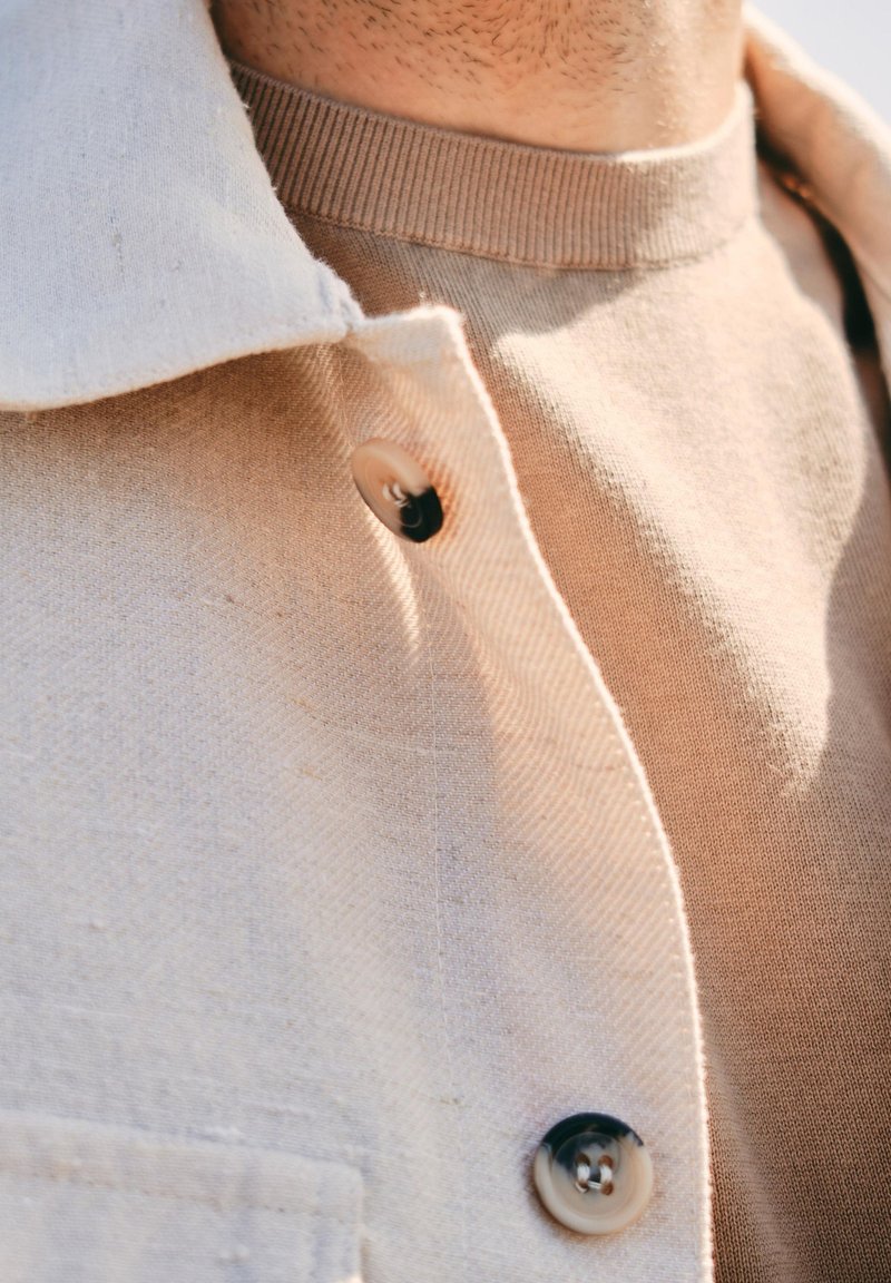 Lichtbeige shirtjack met een gestructureerde stof, met grote knopen in contrasterend zwart. Daaronder is een tan ribgebreide ronde hals trui zichtbaar.