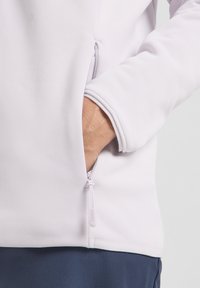 Pullover color lavanda chiaro con texture liscia, dotato di tasca laterale con zip e polsini a coste, presentato senza ulteriori embellimenti.