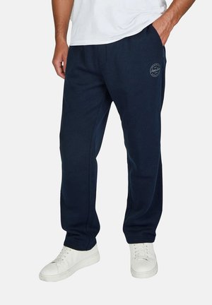 Man wearing navy blue sweatpants with white sneakers and one hand in pocket, standing against a plain background.