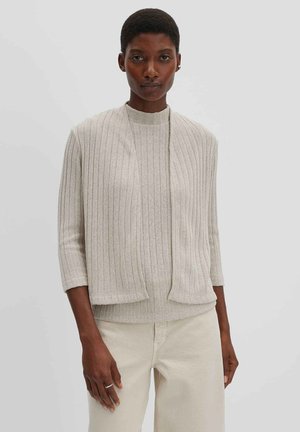 Femme aux cheveux courts portant un pull côtelé beige et un gilet assorti, debout devant un fond gris clair uni.