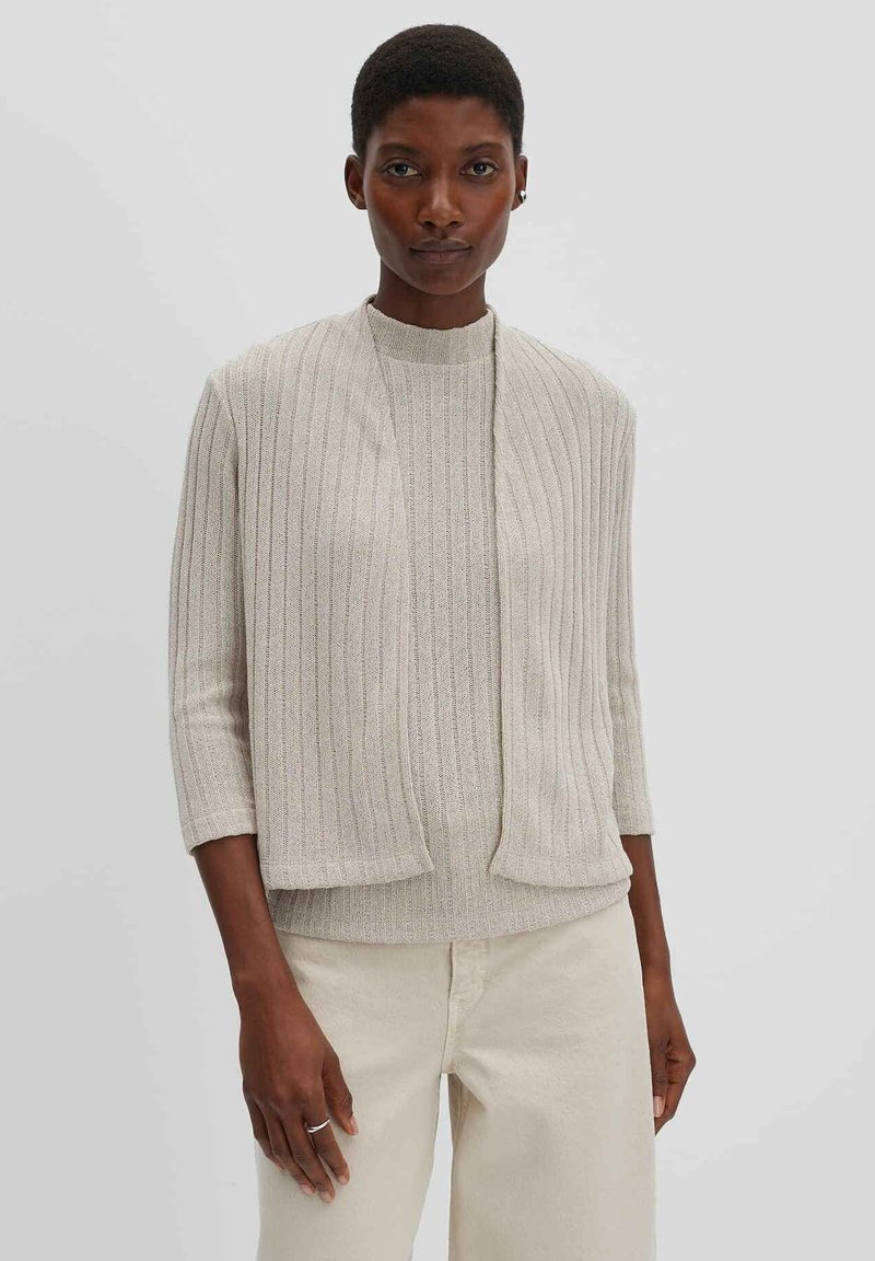 Frau mit kurzen Haaren, die einen beigen gerippten Pullover und eine passende Weste trägt, vor einem schlichten hellgrauen Hintergrund stehend.