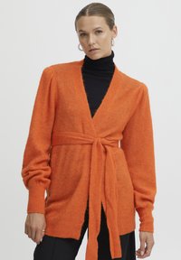 Oranžinė, ilgomis rankovėmis megztinė pūkuotoje tekstūroje su surišamu juosmeniu. Turi gilų v-kaklą ir nerti rankogaliai, derinamas su juoda golfine palaidine.