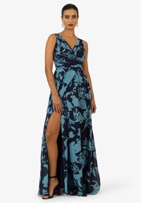 Abito lungo navy con motivo floreale turchese, scollatura a V profonda, corpetto plissè e spacco alto fino alla coscia; abbinato a sandali neri con fasce.