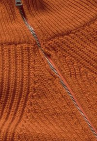 Oranje gebreide trui met een geribbelde structuur, een ritssluiting aan de voorkant en contrasterende zilveren ritsdetails.