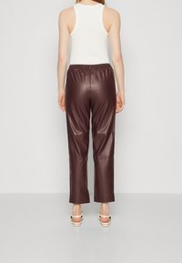 Kobieta ubrana w biały bezrękawnik i wysokie, ciemnobrązowe spodnie ze sztucznej skóry oraz białe sandały na obcasie, stojąca tyłem.