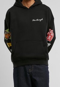 Černá mikina s kapucí a nápisem "Five Boroughs" na hrudi, barevné ovocné a jablečné grafické nášivky na obou rukávech, nošena s tmavými džíny.