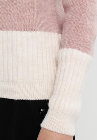 Pull en tricot avec une partie supérieure rose et un ourlet inférieur côtelé crème. Texture douce, manches longues et poignets ajustés qui mettent en valeur les détails du design.