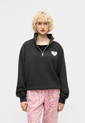 Persona con capelli biondi e ricci che indossa una felpa nera con cerniera a quarti e un emblema a forma di cuore bianco, pantaloni rosa con motivo, in piedi davanti a uno sfondo neutro.