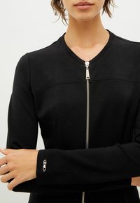 Svart zip-up jacka i mjukt tyg, med rundad krage, långa ärmar och en silverfärgad dragkedja. Logotypdetalj synlig på ärmen.
