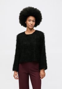 Természetes afro frizurájú nő fekete bolyhos hosszú ujjú felsőt és magas derekú sötétvörös nadrágot visel egy sima fehér háttér előtt.