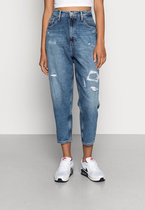 Personne portant un jean bleu taille haute, déchiré, coupé au-dessus des chevilles, avec des baskets Nike blanches et noires et un top blanc court.