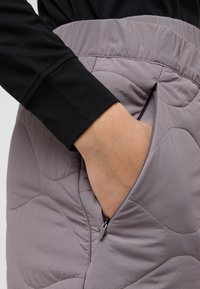 Gequiltede, lavendelrok met een gladde textuur, voorzien van een ritszak aan de zijkant en een elastische tailleband. Zwarte langemouwen shirt zichtbaar.