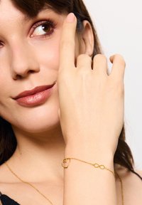 Złota bransoletka z sercem i symbolem nieskończoności, z cienkim łańcuszkiem i delikatnymi ogniwami na nadgarstku ręki, która wskazuje na twarz.