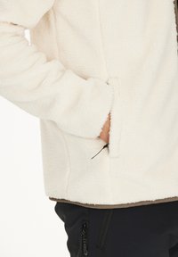 Krämfärgad fleecejacka med en mjuk textur, med en ficka med dragkedja på sidan och elastiska muddar för en tät passform.