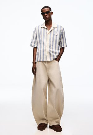 Mężczyzna w czarnych okularach przeciwsłonecznych, niebiesko-białej koszuli w paski z krótkim rękawem, szerokich beżowych spodniach i brązowych butach, stoi z jedną ręką w kieszeni.