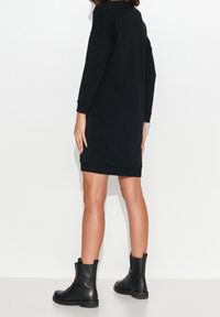 Schwarzes Sweatshirtkleid mit langen Ärmeln und einem Saum, der bis zur Mitte des Oberschenkels reicht. Kombiniert mit schwarzen Schnürstiefeln mit goldenen Hardware-Akzenten.