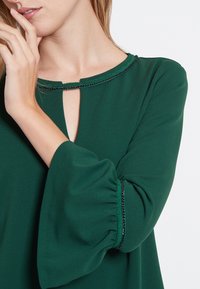 Grön blus i mjukt, flödande tyg. Har en nyckelhålsringning, trumpetärmar och dekorativ kant längs ringningen och  ärmarna.