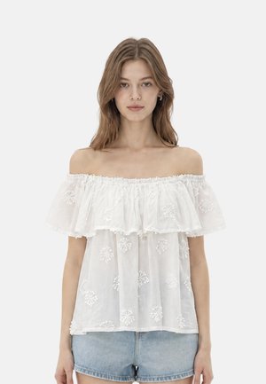 Giovane donna che indossa una blusa bianca con scollo a spalle scoperte e ricami floreali e pantaloncini di jeans azzurri, in piedi davanti a uno sfondo semplice.