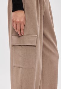 Μπεζ παντελόνι με φαρδιά πόδια, κατασκευασμένο από υφασμάτινη υφή, με μια μεγάλη, ορθογώνια τσέπη cargo στο μηρό και λεπτομέρεια με διπλωμένη ραφή.