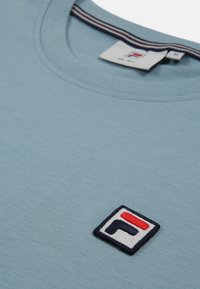 Ljusblå t-shirt med kort ärm och rundad halsringning, prydd med ett strukturämt logopatch i röd, vit och marinblå färg på bröstet.