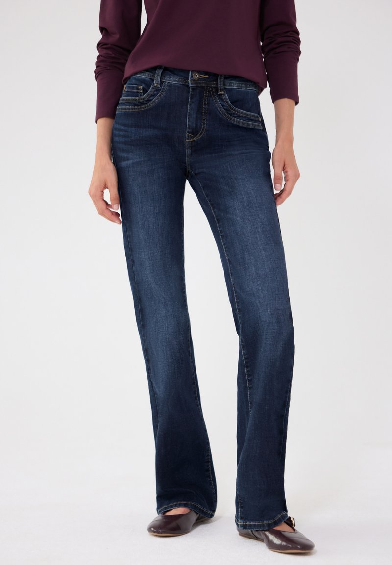 Femme portant un jean bootcut bleu foncé, un haut à manches longues violet foncé et des chaussures plates marron, debout sur un fond blanc.