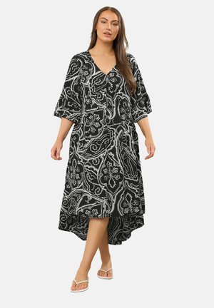 Vrouw draagt een zwart-witte paisleyjurk met V-hals, driekwartmouwen en asymmetrische zoom, gecombineerd met witte slippers.