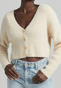 Krémově zbarvený krátký cardigan s výstřihem do V, vyrobený z texturované pleteniny s pěti knoflíky vpředu. Kombinován s světle modrými džínami.