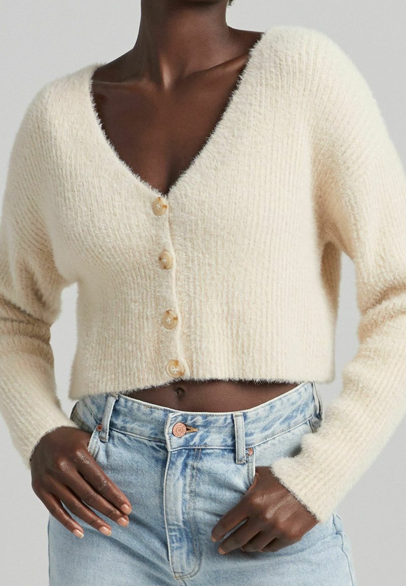 Krémově zbarvený krátký cardigan s výstřihem do V, vyrobený z texturované pleteniny s pěti knoflíky vpředu. Kombinován s světle modrými džínami.