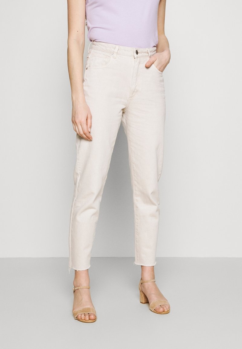 Woman wearing high-waist off-white jeans with frayed hems, a lilac top, and nude block-heeled sandals, standing against a plain background.