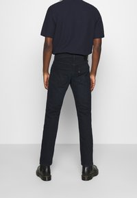 Mörk denimjeans med rak passform, femfickorsdesign, ton-i-ton-sömmar och en subtil logomärke på den bakre midjan. Svarta stövlar synliga.