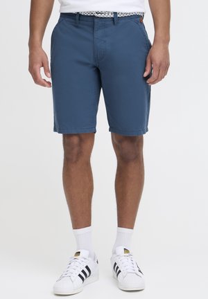 Mand iført blå knælange shorts, hvidt bælte, hvide sokker og hvide Adidas sneakers med sorte striber, stående mod en ensfarvet baggrund.