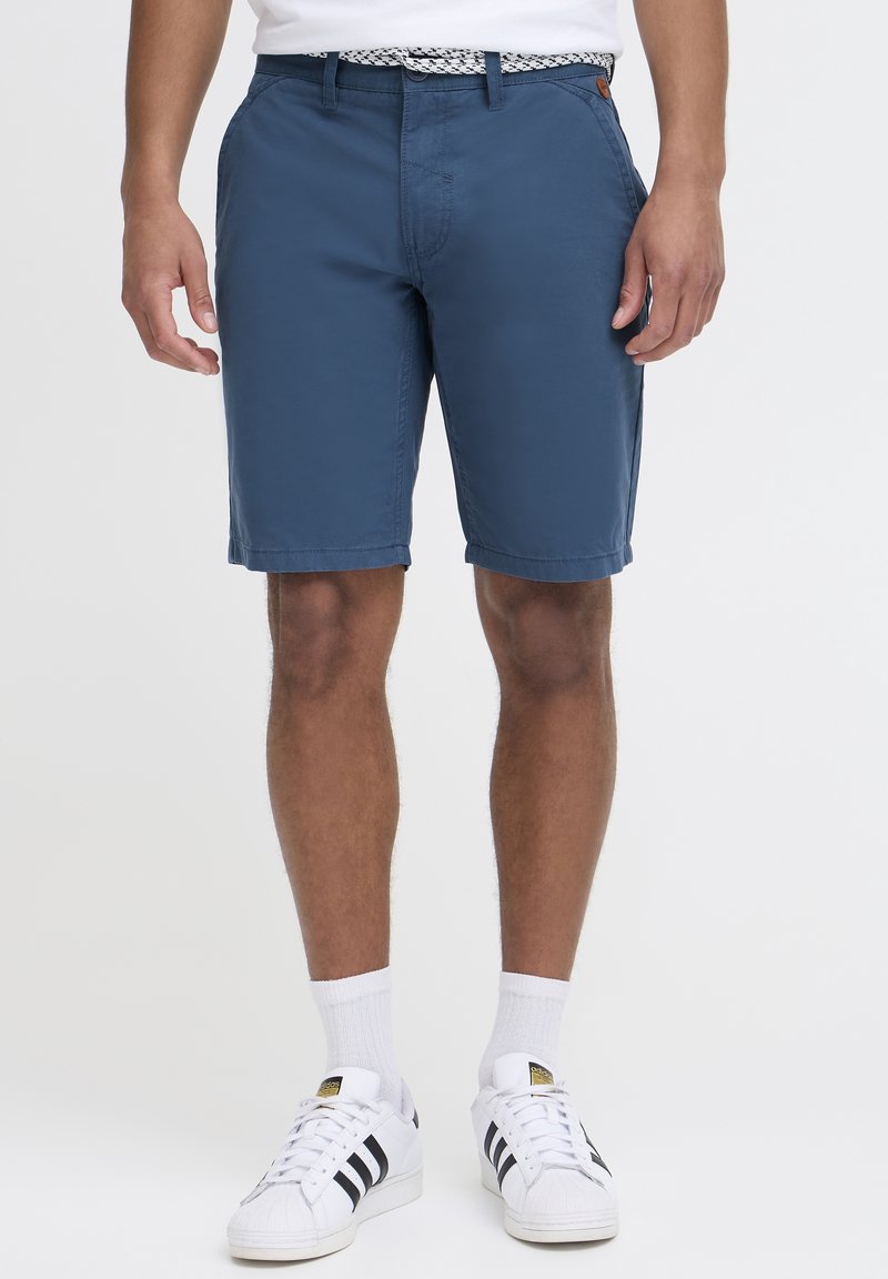 Homme portant un short bleu au genou, ceinture blanche, chaussettes blanches et baskets Adidas blanches à rayures noires, debout devant un fond uni.