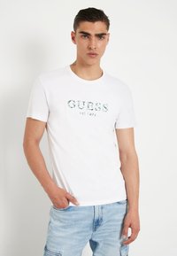 T-shirt en coton blanc à manches courtes, avec "GUESS" imprimé en lettres texturées vertes sur la poitrine, associé à un jean bleu clair.