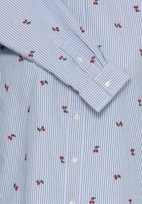 Overhemd met knopen, lichtblauwe en witte verticale strepen, met kleine rode geborduurde kersen en groene bladeren op de stof.