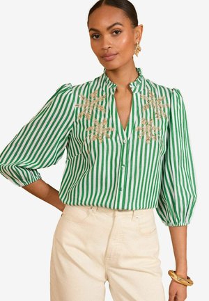 Blusa a righe verde e bianca con colletto alla mandarina, maniche a sbuffo e dettagli ricamati in oro. Abbinata a pantaloni di colore chiaro.